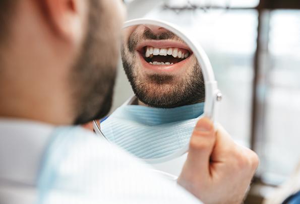 a man looking at his teeth in a handheld mirror in the dental office